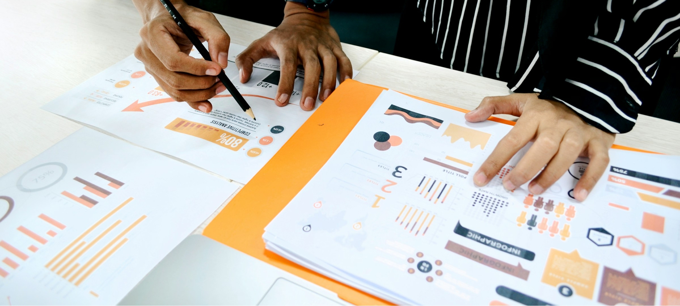 Close-up of two people's hands reviewing printed infographics and charts spread across a desk, with one hand pointing a pencil at a page showing a percentage figure and arrow graphic.