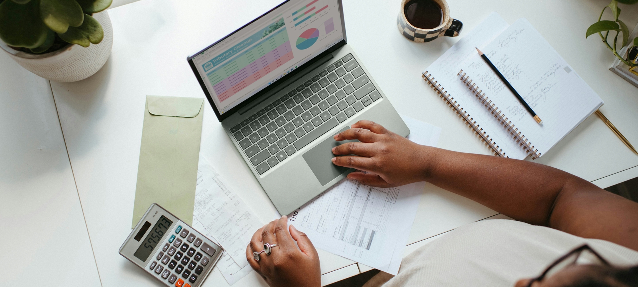 Overhead view of a person's hands using a laptop trackpad, with a financial spreadsheet and pie chart on screen, surrounded by a calculator, printed invoices, an envelope, a notebook, and a cup of coffee on a white desk.